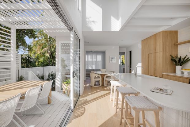 Bright open-plan kitchen and dining area with a large white island, timber cabinetry, light timber flooring, and woven bar stools. Full-height glazed sliding doors open to a covered outdoor dining deck with white slatted screening and outdoor furniture. The interior is filled with natural light and has a clean, contemporary coastal feel.