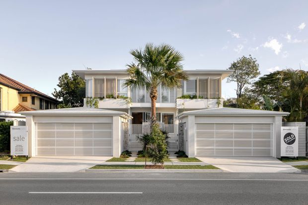 Street-facing exterior photograph of a contemporary white multi-storey residential development with two integrated double garages, a central pedestrian entry, vertical screening, balconies, and a flat roof. Palm trees and landscaped planting frame the facade, with neighbouring homes visible on either side.