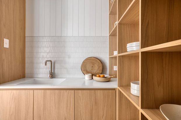 A warm, minimalist kitchen or kitchenette with light timber cabinetry and extensive open shelving. A white integrated benchtop and undermount sink sit beneath a brushed metal tap. The backsplash combines narrow vertical white tiles with vertical white wall panelling above, while simple crockery and wooden serving boards add understated detail.