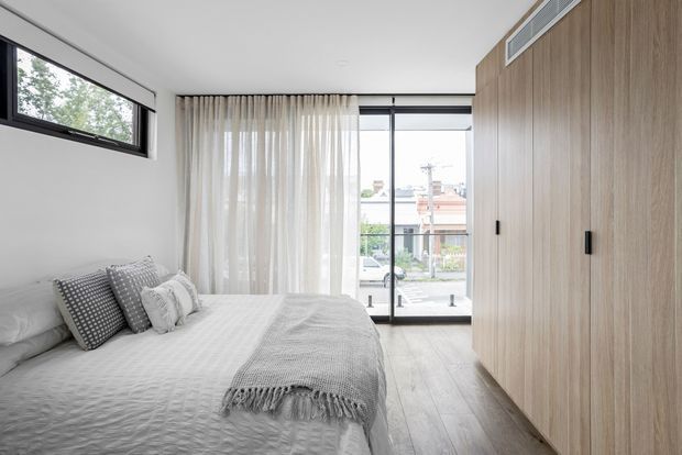 Bright, compact contemporary bedroom with a neatly made bed, layered grey and white bedding, sheer full-height curtains, pale timber flooring, and a full-height light timber built-in wardrobe. A large glazed door or window opens to an urban street view, while a high horizontal window brings in additional natural light. The room has a clean, restrained and airy feel.