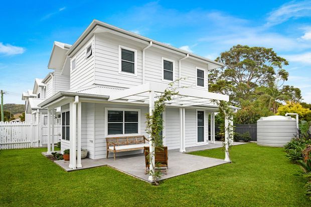 Two-storey white weatherboard house with a pitched roof, white-framed windows, covered outdoor patio, pergola with climbing plants, manicured lawn, picket fencing and a large rainwater tank. The exterior has a bright coastal-residential character.