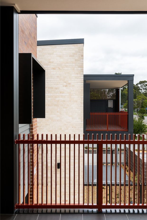 View from a covered upper-level or balcony walkway toward a compact contemporary residential facade. The building features pale exposed brick walls, darker brickwork, charcoal metal roof and trim elements, black vertical screening, and red-brown metal balustrades and fencing. A neighbouring covered outdoor entry or carport-like structure is visible beyond, with suburban greenery in the background.