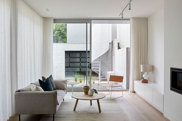 Bright contemporary living room with a neutral upholstered sofa, round timber coffee table, pale rug, white built-in media cabinetry, fireplace, floor-to-ceiling curtains, and large glazed doors opening to a modern courtyard. A sculptural spiral staircase and white masonry walls are visible beyond the glazing.