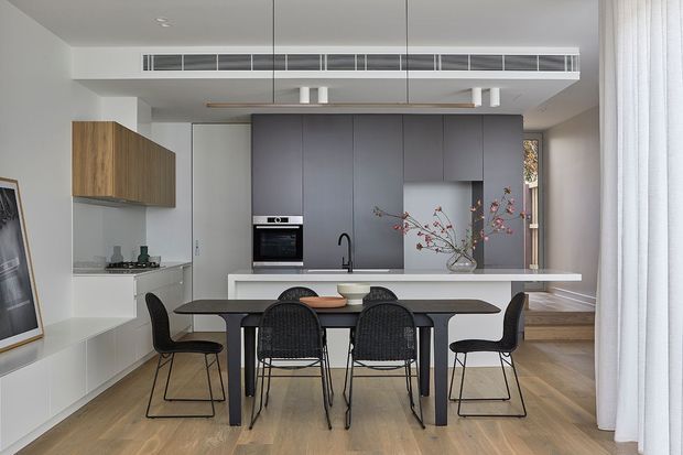 A contemporary open-plan kitchen and dining area with a long island bench, integrated dark cabinetry, timber upper cabinets, and a black dining table surrounded by woven and upholstered chairs. Pale timber flooring, white walls, sheer curtains, and linear ceiling ventilation create a clean, refined interior. A large window or glazed opening brings natural light into the space.