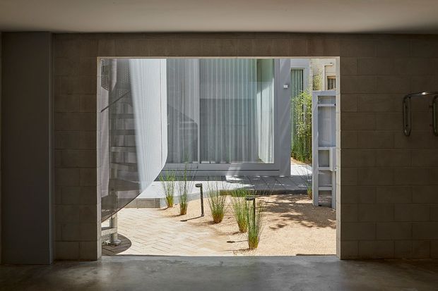 View from a dark, minimally finished interior or covered space through a large open doorway into a landscaped courtyard. The opening is framed by large rectangular grey wall tiles, with a smooth concrete floor and exposed door hardware. Outside, pale paving, gravel, ornamental grasses, and a contemporary glazed building façade are visible.