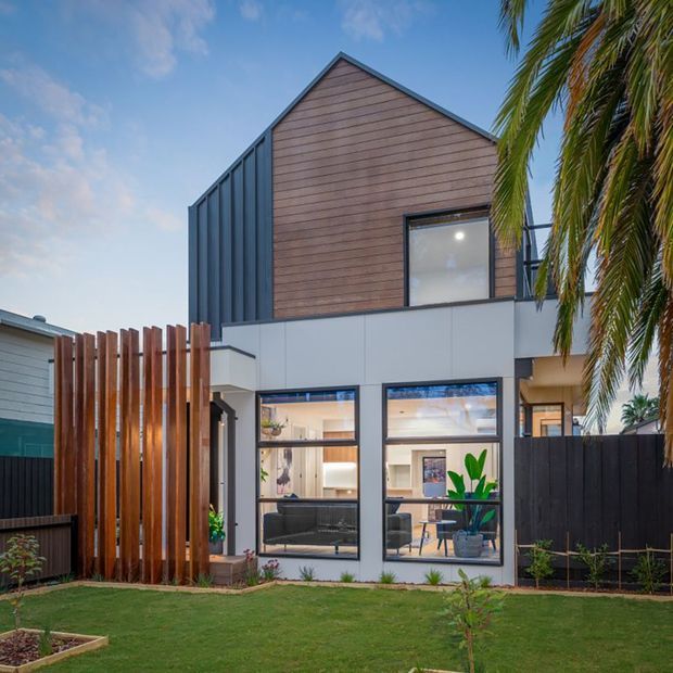 Exterior view of a contemporary multi-level house with a steep gabled form, warm horizontal timber-look cladding, dark vertical metal cladding, pale panelled lower facade, and large ground-floor windows. A tall timber-slat privacy screen, dark fencing, lawn, and tropical palms frame the frontage.