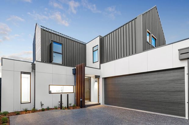 Contemporary multi-level residential facade viewed from the driveway, featuring crisp white wall panels, dark vertically ribbed metal cladding, angular pitched roof forms, narrow blue-tinted windows, a timber entry screen, and a wide integrated double garage. The forecourt is finished in dark aggregate paving with minimal landscaped planting.