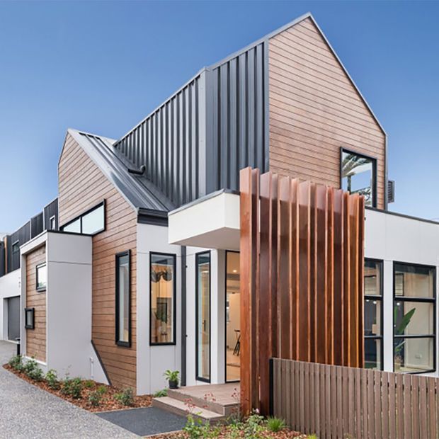 Exterior view of a contemporary multi-level residence with interlocking gabled volumes, pale horizontal timber cladding, dark corrugated metal roofing and wall panels, white rendered sections, black-framed glazing, and a prominent vertical timber privacy screen at the entry. The home sits beside a gravel driveway and landscaped planting.