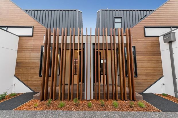 Front exterior view of a contemporary residential building with symmetrical timber-clad wings and dark grey standing-seam metal roof forms. A central recessed entry is screened by vertical timber and metal battens, with white angular wall panels, narrow windows, landscaped gravel beds, and dark paved paths.