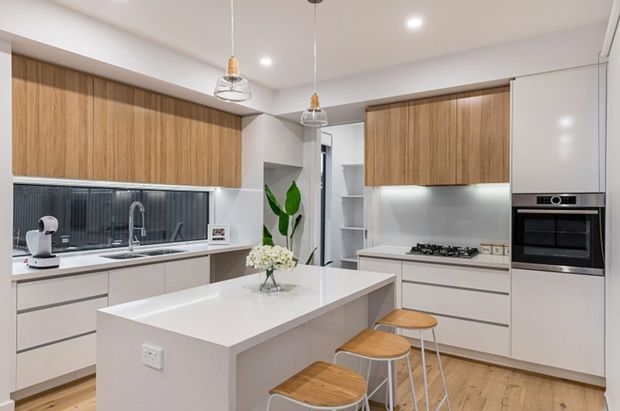 Bright contemporary white-and-timber kitchen with a large central island, integrated cabinetry, timber stools, pendant lights, stainless steel sink and tap, gas cooktop, and built-in oven. The space has light timber flooring, white cabinetry and benchtops, and a dark textured splashback behind the sink.