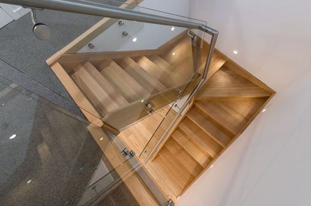 View looking down into a modern stairwell with warm timber stairs and landing, clear glass balustrades, and sleek metal handrails. Recessed lighting highlights the timber construction, while dark grey tiled flooring and white walls create a clean contemporary setting.