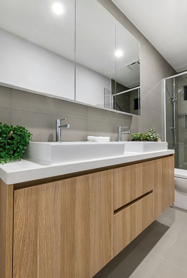 Contemporary bathroom featuring a floating double vanity with light timber cabinetry, a white solid-surface benchtop, two square white basins, chrome tapware, a large frameless mirror, and pale grey wall and floor tiles. A glass-enclosed shower and part of a bathtub are visible at the right. Small green plants add natural accents.