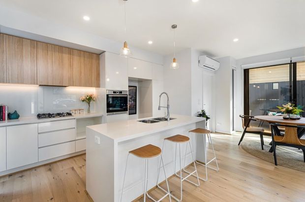 Bright, open-plan contemporary kitchen and dining area with white flat-panel cabinetry, warm timber overhead cupboards, a large white island with integrated sink and four timber-seated stools, stainless appliances, and pale timber flooring. Pendant lights and recessed ceiling lights illuminate the space, while full-height glazing opens to an outdoor area beyond the dining zone.