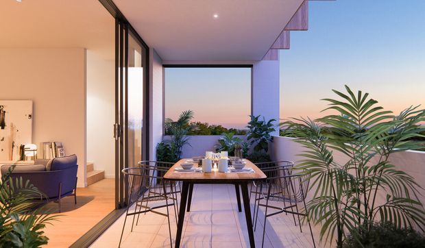 A contemporary apartment dining terrace with a rectangular outdoor dining table and wire-frame chairs, surrounded by lush potted palms and plants. Full-height black-framed glazing opens onto the adjacent living area, while a covered ceiling and balcony parapet frame views of a pastel sunset skyline. The warm interior lighting and pale finishes create an inviting indoor-outdoor setting.