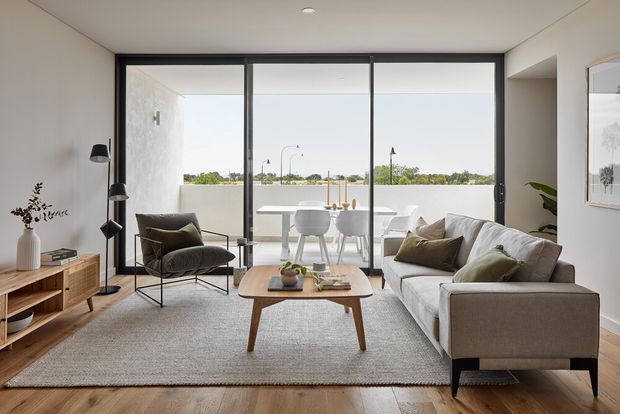 Bright contemporary living room with a neutral palette, timber flooring, a large textured area rug, low timber media console, sculptural floor lamp, armchair, coffee table, and a pale upholstered sofa. Full-height black-framed glazing opens the room visually to a balcony or terrace and distant greenery, with a dining area visible beyond.
