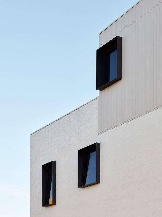A close-up exterior view of a minimalist contemporary building facade. The composition shows clean geometric volumes clad in pale white materials, with three dark-framed windows projecting from the wall and contrasting strongly against the light facade. The image is tightly cropped against a clear blue sky, emphasizing the angular roofline and modern architectural detailing.