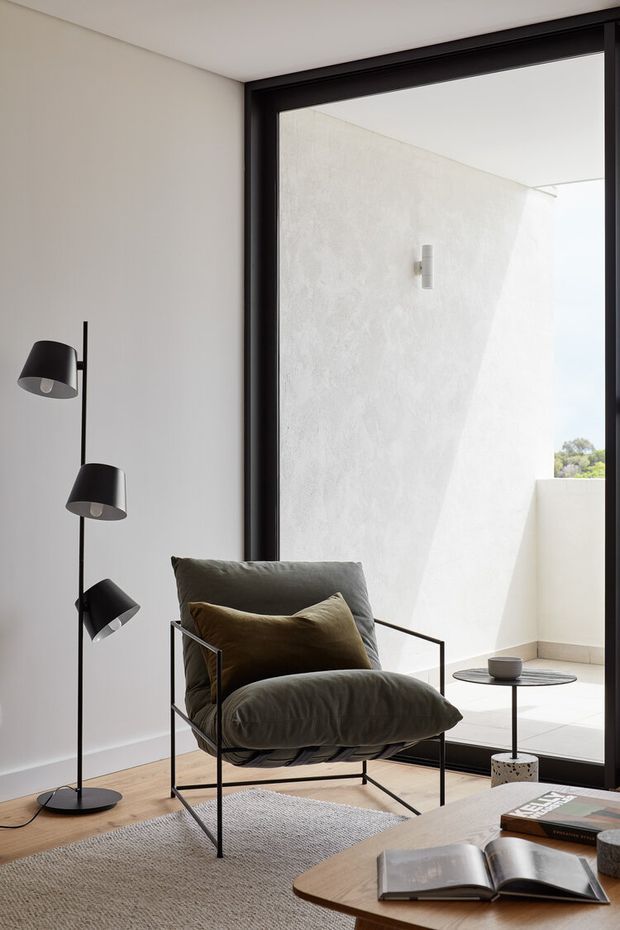 A refined contemporary living room corner with a black metal-framed armchair, charcoal floor lamp, small side table, and low timber coffee table with books. Neutral plaster walls, pale timber flooring, a textured light rug, and extensive black-framed glazing create a bright minimalist composition. The glazing opens toward a small white-walled terrace or courtyard.