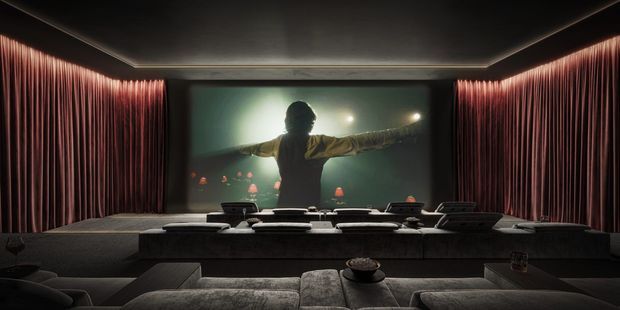A dark, luxurious home cinema with tiered rows of deep upholstered seating facing a large projection screen. Floor-to-ceiling burgundy curtains frame the screen, while recessed perimeter lighting and a low dark ceiling create an immersive theatrical atmosphere. A person is visible on the projected image.