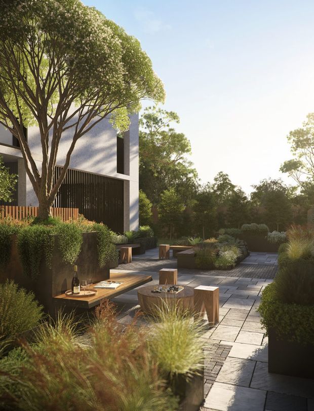 A landscaped outdoor courtyard or terrace beside a contemporary multi-storey building. The space features large-format grey paving, raised dark planter beds, clipped shrubs, ornamental grasses, mature trees, built-in bench seating, and small timber stools and tables arranged for outdoor gathering. The building has a restrained modern facade with dark vertical screening and light rendered or concrete walls. Warm low-angle sunlight creates a refined, tranquil garden atmosphere.