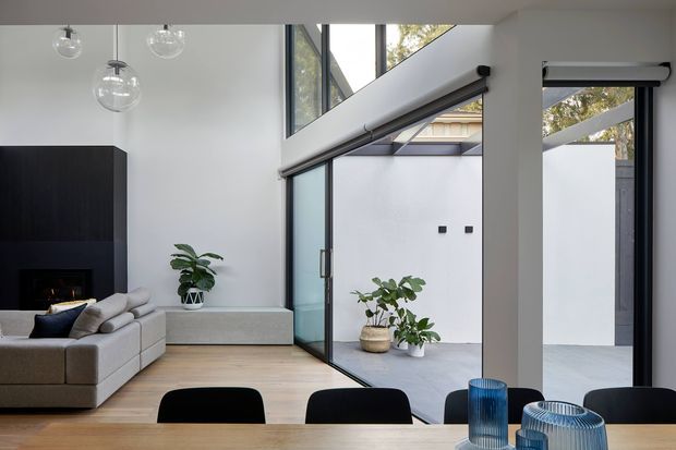 A bright contemporary open-plan living and dining area with a large light-grey sectional sofa, black built-in fireplace wall, timber flooring, and a simple timber dining table with black chairs. Floor-to-ceiling glazed doors and windows open toward a small paved courtyard, with indoor plants and sculptural glass pendant lights adding detail. The space has a tall, airy ceiling and strong indoor-outdoor connection.
