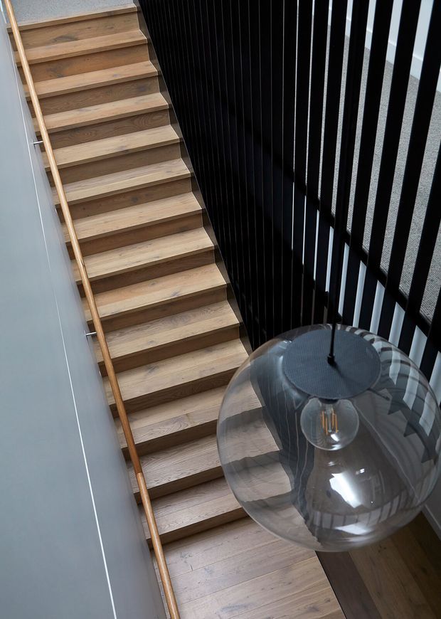 Interior view looking down a timber staircase with open wooden treads, a slim timber handrail, and a dramatic black vertical slatted screen along one side. A large smoked-glass pendant light hangs in the stairwell, with pale walls and timber flooring contributing to a refined contemporary composition.