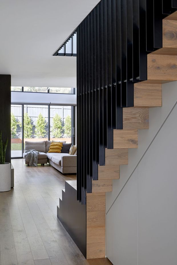 Interior view dominated by a sculptural open staircase with floating natural timber treads and a striking black vertical slatted balustrade or screen. The staircase sits beside a bright open-plan living area with pale timber flooring, floor-to-ceiling glazing, a neutral sectional sofa, and a yellow accent cushion. The space has clean contemporary detailing, strong geometric lines, and abundant daylight.