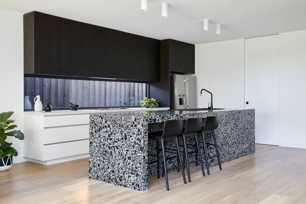 Contemporary kitchen with a large waterfall island clad in a bold black-and-white terrazzo-like surface, integrated dark timber cabinetry, white lower cabinets, a dark textured splashback, and four black bar stools. The bright open space has pale timber flooring, white walls and ceiling, minimalist cylindrical downlights, a black tap, and concealed full-height storage or appliance cabinetry.