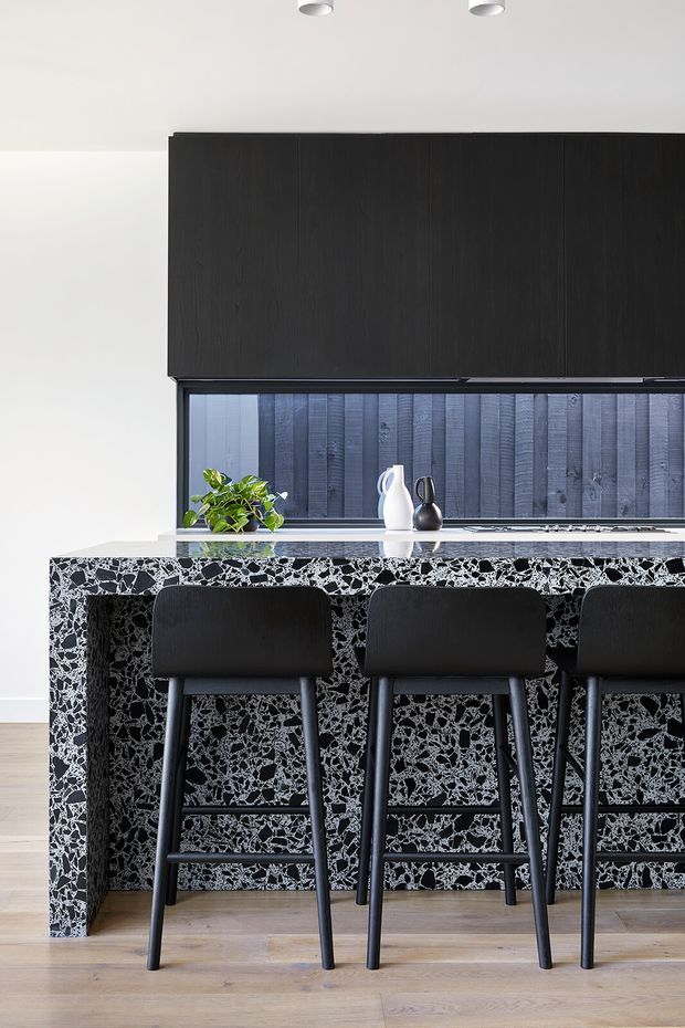 A contemporary kitchen with a dramatic black-and-white terrazzo island and matching splashback. The island includes seating for three at slim black upholstered stools, while dark timber-grain overhead cabinetry contrasts with white walls and pale timber flooring. A small plant and monochrome decorative vessels sit on the benchtop, with recessed ceiling spotlights above.