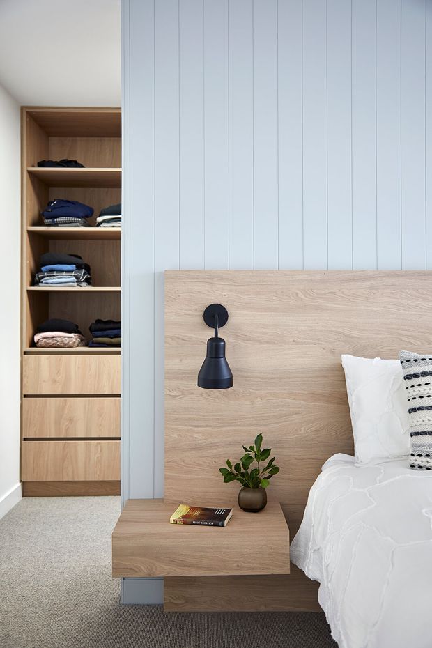 A bright, refined bedroom with a built-in open wardrobe and drawers, pale vertical wall panelling, a light timber headboard, and a floating timber bedside table. The bed is dressed in white bedding with a patterned cushion, complemented by a matte black wall sconce, a small potted plant, and neutral carpet.