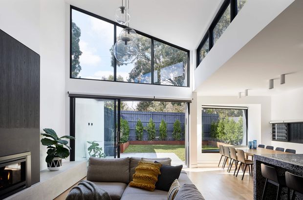 Bright, open-plan contemporary living area with a double-height ceiling and extensive black-framed glazing overlooking a landscaped courtyard. The room combines a grey sectional sofa, suspended glass globe pendant lights, a dark feature wall with fireplace, and a stone kitchen island with seating. A dining area is visible beyond the living space, with a mezzanine-like upper-level opening and clerestory windows contributing to the airy volume.