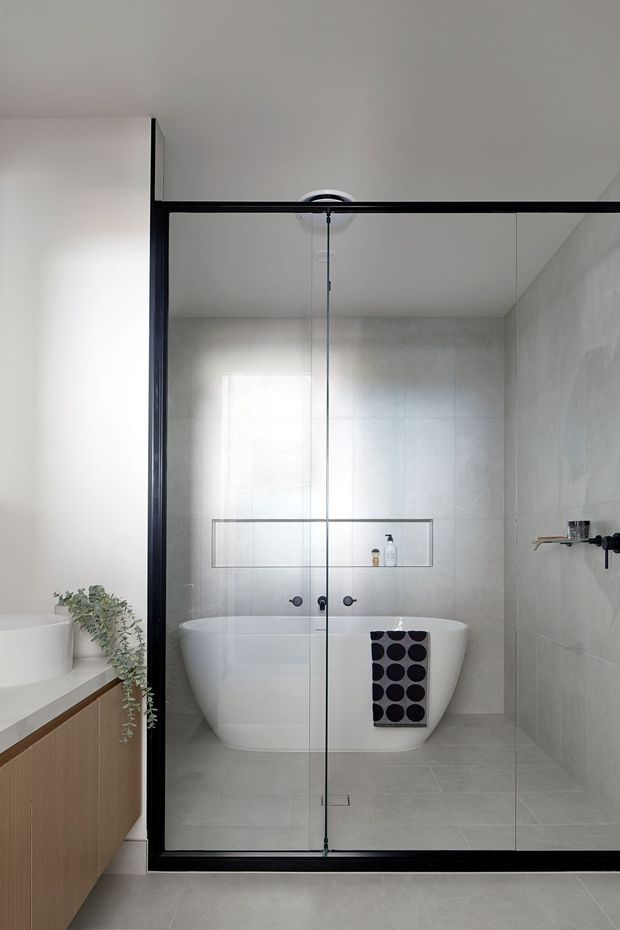 Contemporary bathroom featuring a freestanding white bathtub enclosed behind a black-framed frameless glass shower screen. Large pale grey wall and floor tiles create a clean, minimal backdrop, with a recessed shelf, wall-mounted black fixtures, and a timber vanity with a white basin visible at left. A trailing plant and patterned towel add subtle contrast.