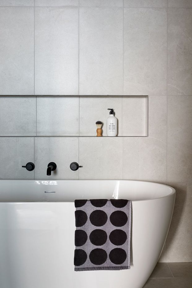 Contemporary minimalist bathroom featuring a freestanding white bathtub, large-format pale grey stone-look wall tiles, dark wall-mounted bath fixtures, and a recessed shelf with soap and a brush. A monochrome patterned towel adds contrast.