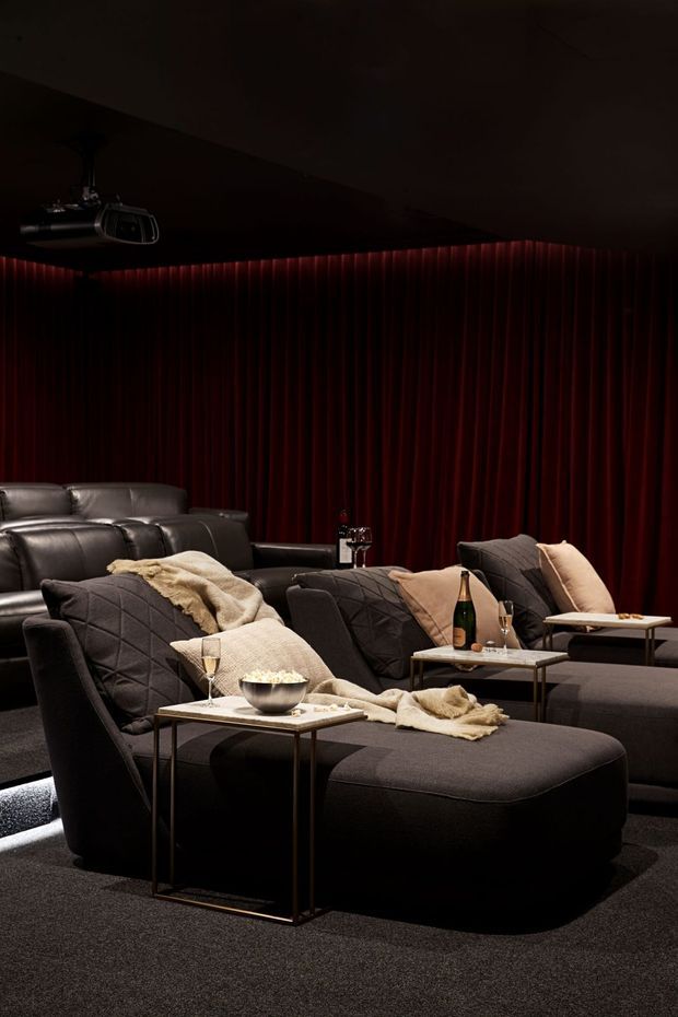 A dark, luxurious home cinema with multiple large charcoal sectional recliner seats and ottomans arranged in rows. Deep burgundy velvet curtains cover the walls, while small side tables hold drinks, snacks and champagne. The room has a fully carpeted floor, concealed low lighting, and a ceiling-mounted projector, creating an intimate theatrical atmosphere.