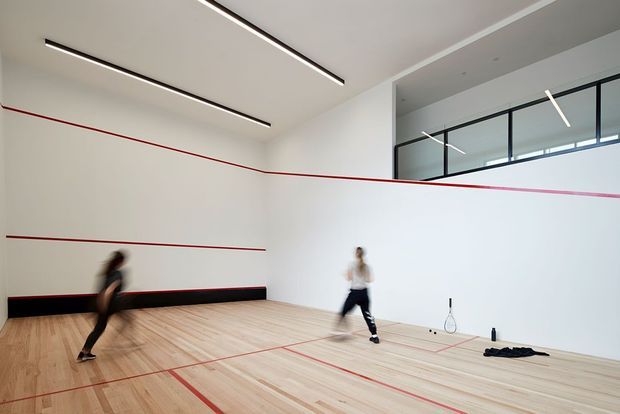 Bright modern indoor squash court with pale timber flooring, white walls, red court markings, black skirting, linear ceiling lights, and a glazed upper viewing area. Two players are captured in motion, with squash rackets and equipment on the floor.