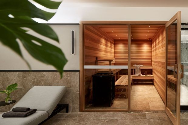 A luxurious indoor sauna suite viewed from a spa-like relaxation area. The sauna has warm timber wall and ceiling lining, tiered timber benches, a central sauna heater, and a glass door and partition. The surrounding room features large-format stone-look flooring and wall cladding, a padded treatment or relaxation bench, soft neutral finishes, and tropical greenery.