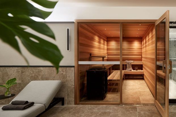 A refined home sauna and relaxation area. The image shows a timber-lined sauna cabin with a glazed timber door, tiered timber benches, warm integrated ceiling lighting, and a dark sauna heater. In the foreground is a padded treatment or relaxation bench beside a large indoor tropical plant. The surrounding room has stone-look wall and floor finishes and a calm, spa-like atmosphere.