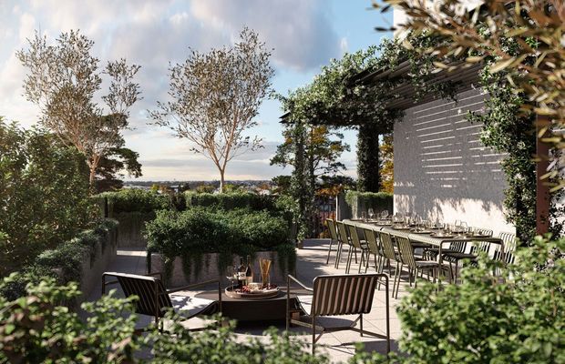 A landscaped outdoor terrace with a long dining table and chairs beside a contemporary building. Trimmed hedges, planter boxes, mature trees, climbing greenery, and a separate lounge arrangement create a refined garden setting. The terrace uses light paving and dark timber or metal architectural screens, with distant landscape views beyond the garden.