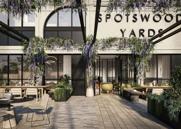 A contemporary landscaped courtyard and outdoor dining area at Spotswood Yards. The image shows a multi-storey building facade with large black-framed windows, arched glazing, pale textured walls, a prominent site sign, timber-look decking, climbing wisteria, raised planters, outdoor tables and chairs, and a glazed entrance leading indoors.