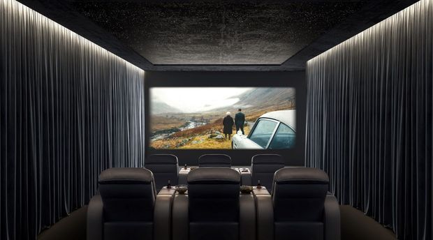 A dark, dedicated home cinema with three rows of black reclining cinema seats facing a large projection screen. The room has floor-to-ceiling vertically ribbed wall panels, a textured dark ceiling, and concealed linear lighting along the upper side edges. The screen displays a landscape scene, creating a dramatic, immersive viewing environment.