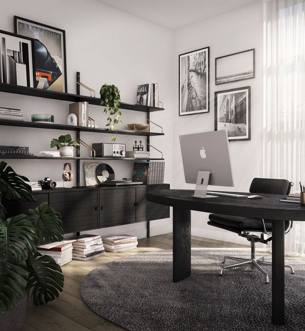A stylish home office with a dark timber desk, computer monitor, ergonomic office chair, and extensive black open shelving with integrated low cabinetry. The shelves display books, framed artwork, audio equipment, decorative objects, and indoor plants. White walls, monochrome photography, sheer floor-to-ceiling curtains, timber flooring, and a large round textured rug create a refined contemporary monochrome scheme.