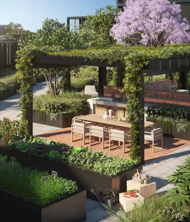 A landscaped outdoor dining and entertaining area beneath a dark timber pergola heavily covered in climbing greenery. The space includes a long timber dining table with multiple chairs, raised planter beds filled with vegetables and ornamental grasses, paved walkways, built-in garden joinery, and a partially visible contemporary building behind dense planting. A flowering purple tree forms a prominent backdrop.