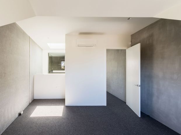 A compact, minimalist interior corridor or room with dark grey carpet, smooth grey textured walls, white ceiling and built-in white partitions. An open white door leads into an adjoining space, while a bright opening at the rear reveals a partially visible bathroom or wash area with a mirror. The room is sparsely furnished and receives strong natural light.