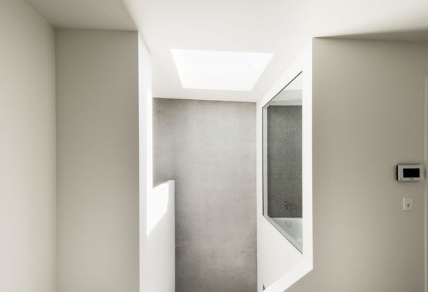 Minimal, bright interior view of a hallway or circulation space leading toward a partially visible bathroom with a glass shower enclosure. White walls and ceiling surround a grey concrete-look wall, with a large skylight providing natural illumination. A small wall-mounted control panel is visible on the right.