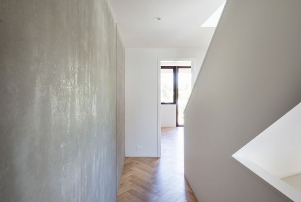 A bright minimalist interior hallway or landing with a dramatic sloping stair enclosure on the right, a textured grey feature wall on the left, and warm herringbone timber flooring. Natural light enters through a skylight and tall dark-framed window at the end of the passage.