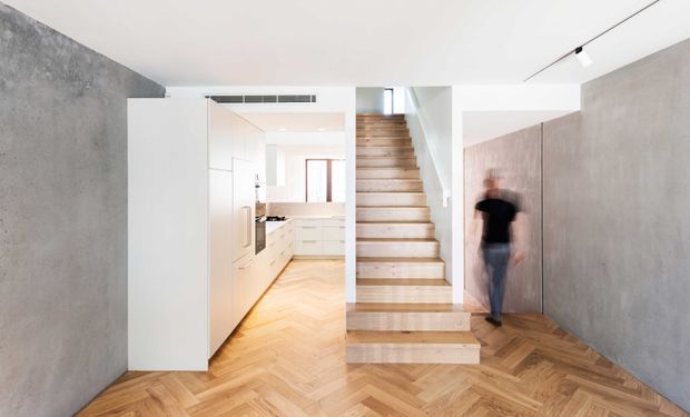 Bright contemporary interior showing an open-plan entry and circulation area with a central timber staircase, herringbone timber flooring, white built-in kitchen cabinetry in the background, and smooth grey concrete-look walls. Minimal linear track lighting and clean architectural detailing create a refined, modern feel.