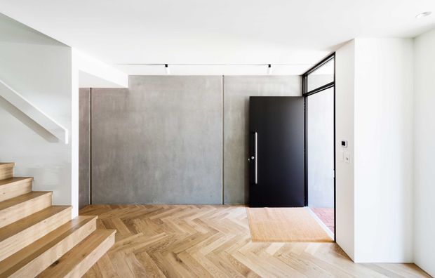 Bright contemporary entryway with a black pivot front door, full-height grey concrete-look feature wall, white walls and ceiling, and warm herringbone timber flooring. A timber staircase rises along the left side, while a slim black-framed glazed panel sits beside the entrance. Minimal track lighting and restrained detailing create a clean, modern foyer.