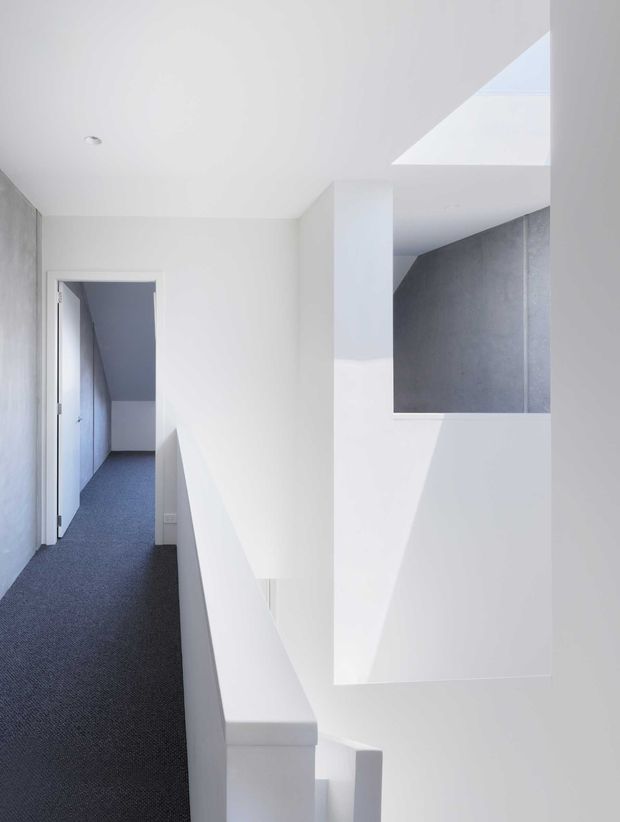 A bright minimalist interior view of a narrow upper-level hallway beside an open stairwell. White walls, ceiling and stair balustrades create a clean geometric composition, while dark grey carpet runs along the corridor. A grey textured wall surface is visible across the stair void, with strong natural light entering from an upper opening or skylight.