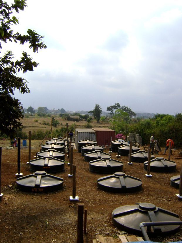 Outdoor rural installation site with numerous black cylindrical water tanks arranged across bare earth, each fitted with a raised circular access lid and connected or supported by vertical posts. A few workers and storage containers are visible in the background, with open farmland and trees beyond under an overcast sky.