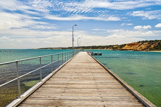 A timber pier or jetty extends over clear turquoise coastal water, with simple metal safety railings and slim lamp posts along the walkway. A sandy, vegetated shoreline and low rocky cliffs are visible in the distance beneath a bright blue sky with scattered clouds.