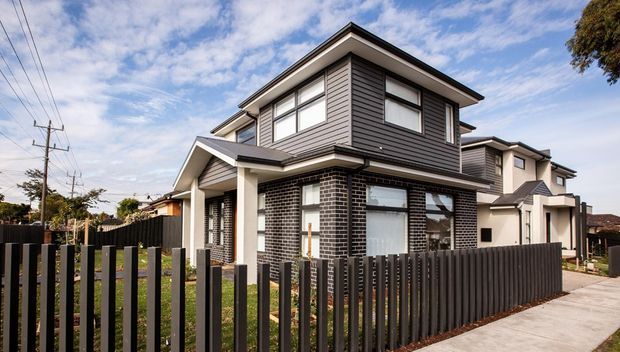 Exterior view of a contemporary multi-level residential townhouse with dark brickwork on the ground floor, grey horizontal weatherboard cladding above, a low-pitched roof, white-framed windows, and a dark vertical-slat front fence. A paved driveway and neighbouring attached dwellings are visible.
